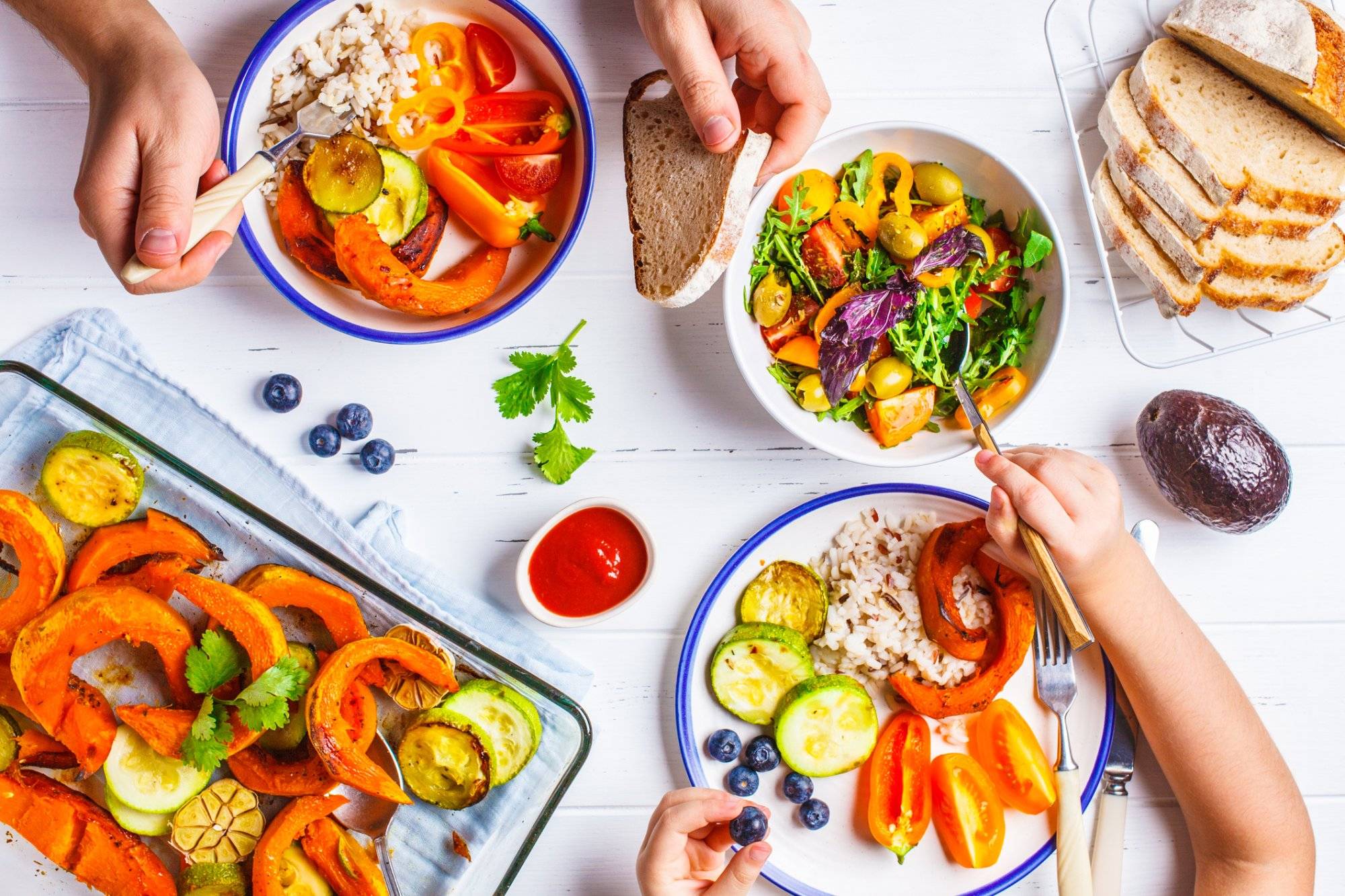 Family eating a healthy vegetarian food. Vegan lunch table top view, plant based diet. Baked vegetables, fresh salad, berries, bread on a white background. Family eating a healthy vegetarian food. Vegan lunch table top view, plant based diet. Baked vegetables, fresh salad, berries, bread on a white background.